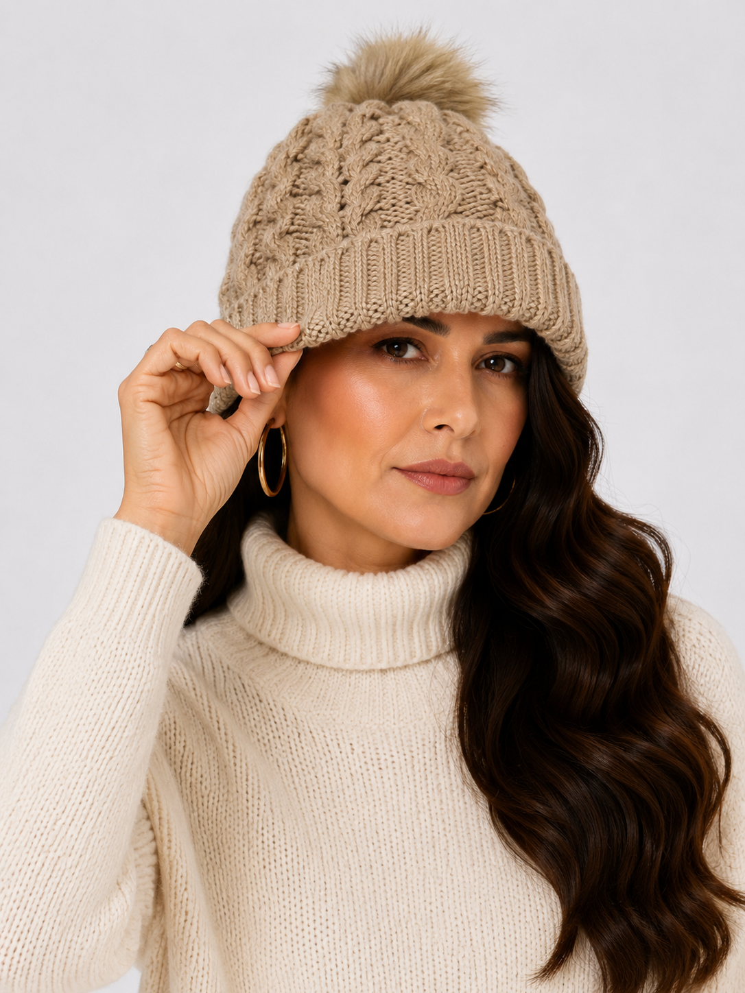 Mujer usando gorro tejido beige con diseño de trenzas y pompón de pelo sintético.